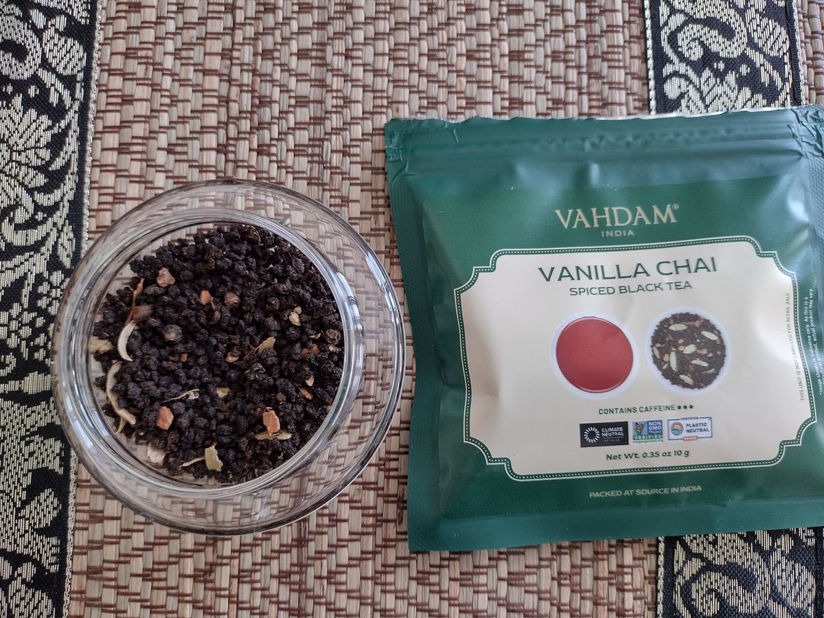 Top-down close-up of the dry Vanilla Chai blend in a clear jar. Small, dark brown CTC tea granules are mixed with visible pieces of lighter spices (like cardamom pods or ginger). The green Vahdam Vanilla Chai packet lies next to the jar on a textured, woven placemat.