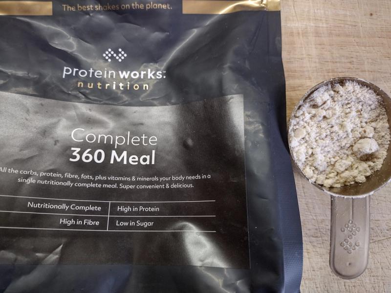     Query successful  A scoop filled with a beige-coloured powder rests next to a black bag of Protein Works Nutrition Complete 360 Meal powder on a light wooden surface. The bag's label highlights that the product is a "nutritionally complete" meal replacement that is "high in protein" and "high in fibre." The top of the bag has the text "The best shakes on the planet.