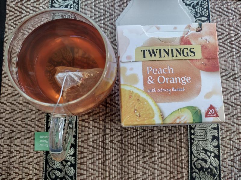 A top-down shot of a Twinings Peach & Orange infusion box next to a clear glass mug of the brewed tea, with the pyramid teabag visible inside.