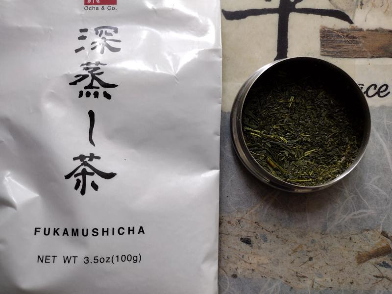 Overhead shot of Ocha & Co. Fukamushicha packaging with Japanese characters for 'Fukamushicha' (深蒸し茶). To the right, an open container reveals the fine, deep green loose leaf tea. The background is a textured, artistic surface."