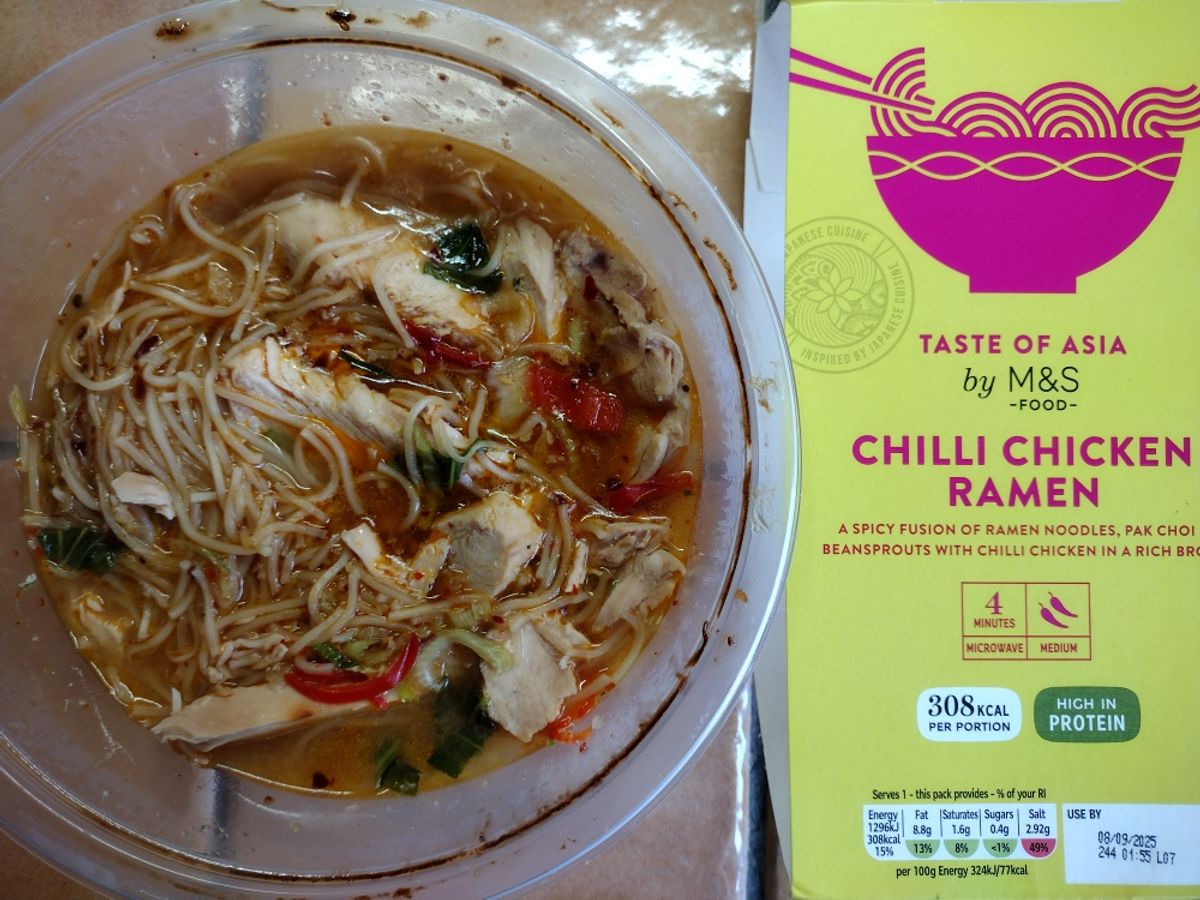 An overhead review photo of the M&S Taste of Asia Chilli Chicken Ramen. The prepared noodle soup is shown in its bowl next to the distinctive yellow and pink product packaging.