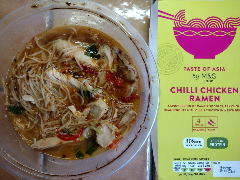 An overhead review photo of the M&S Taste of Asia Chilli Chicken Ramen. The prepared noodle soup is shown in its bowl next to the distinctive yellow and pink product packaging.