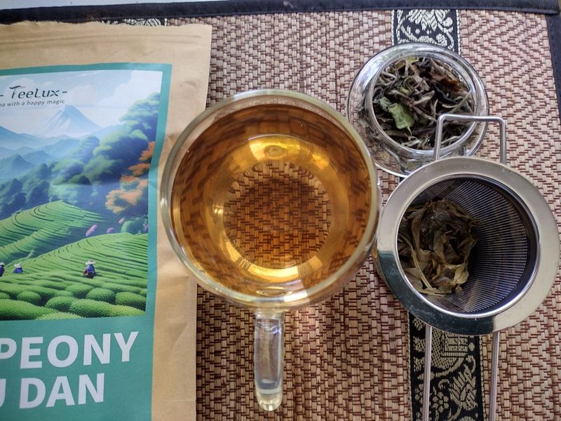A prepared cup of Teelux White Peony tea, showing the golden liquor, alongside the infused Bai Mudan loose leaves in a strainer and the original packaging on a woven mat.