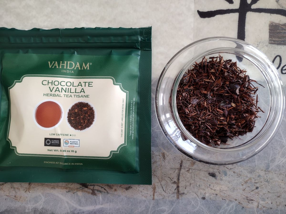 Overhead close-up of the dry Chocolate Vanilla tisane blend in a clear jar. The mix consists primarily of fine, reddish-brown Rooibos needles interspersed with darker, coarser fragments likely from cocoa husks or nibs. The Vahdam packaging is placed above the jar.