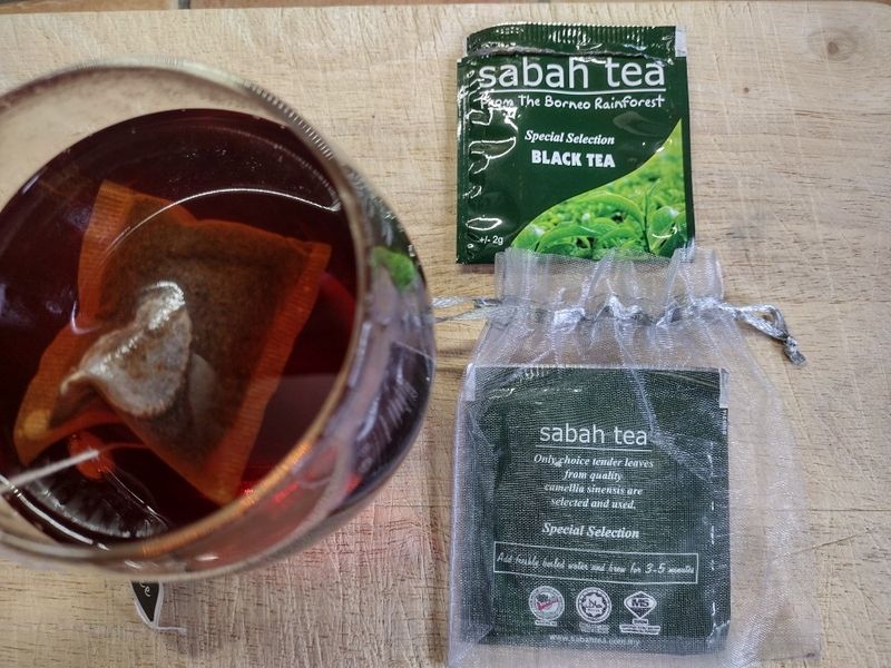 Tasting experience of organic Sabah Tea, showing the dark brewed liquor in a glass, the tea bag, and the vibrant green sachet from the single-estate Borneo plantation