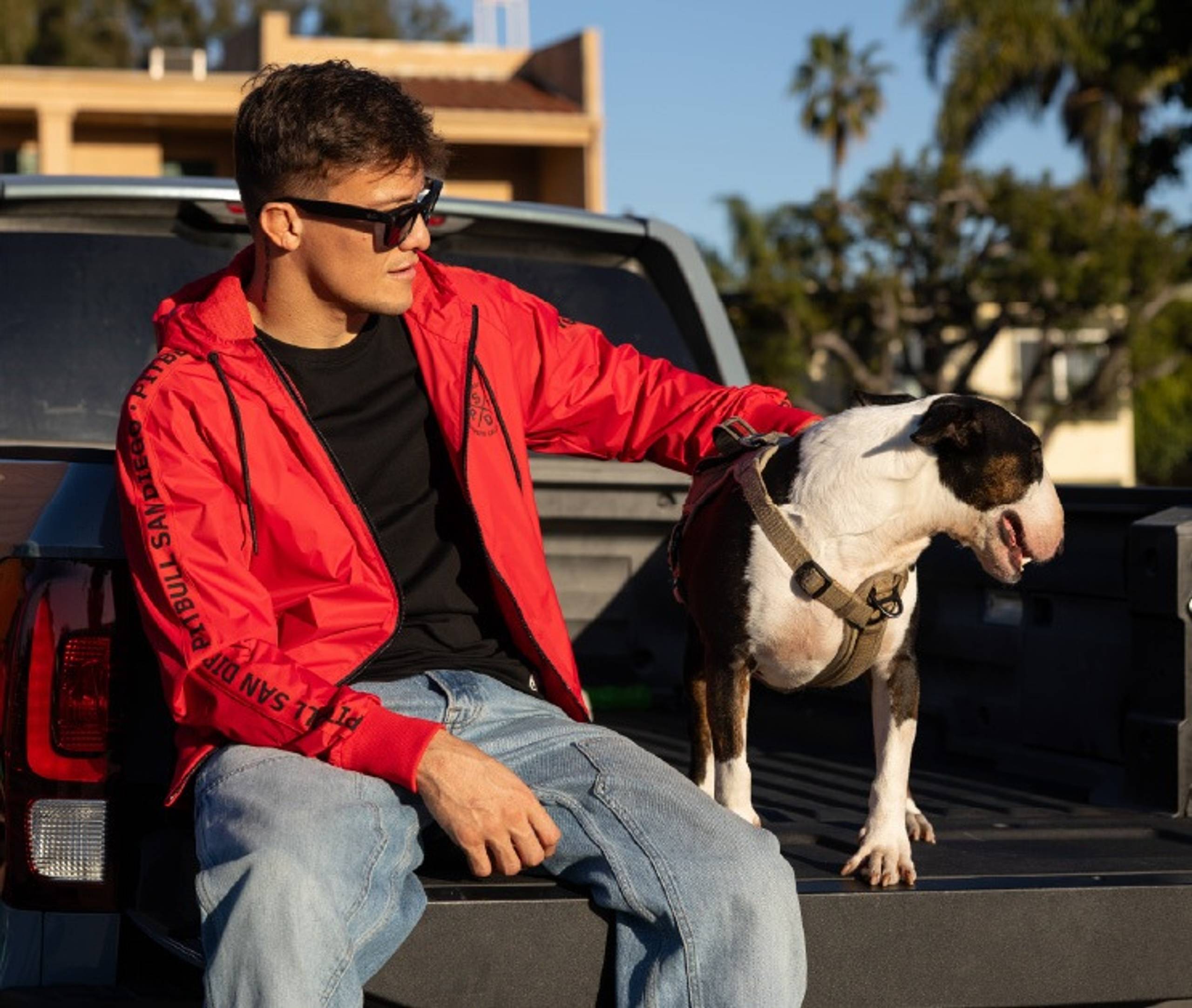 Man in red Pitbull jacket petting a dog in a truck bed