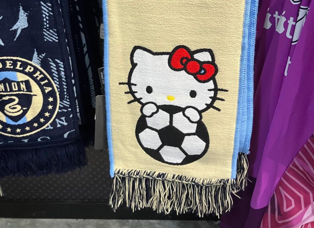 In the center of the image is a yellow scarf with a blue lining with an image of Hello Kitty. It shows her white head and iconic red bow as she holds a soccer ball.