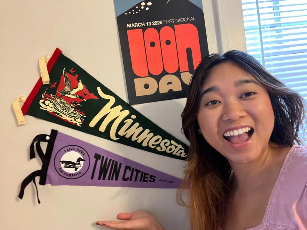 Woman smiles at camera and holds hand up as if to display two pennant flags hanging on wall behind her