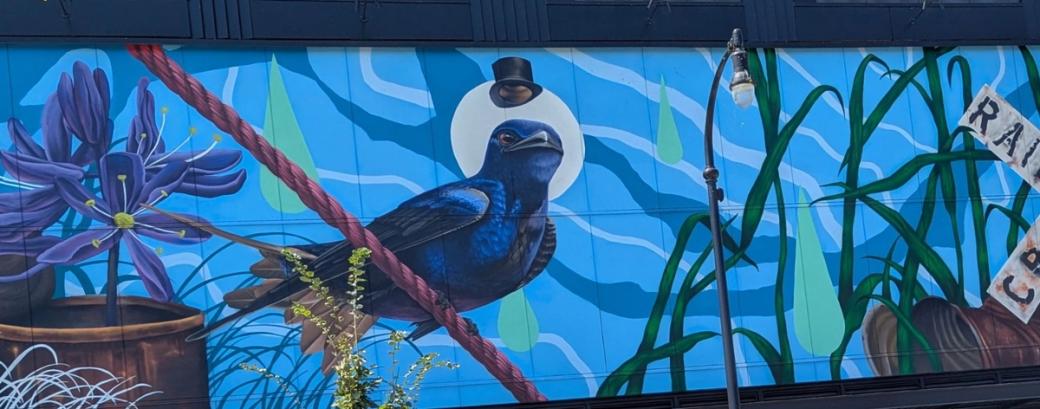 A painted mural with purple and yellow flowers to the left, a blue bird with a top hat floating above its head, and thin green plants to the right with a blue and white background.