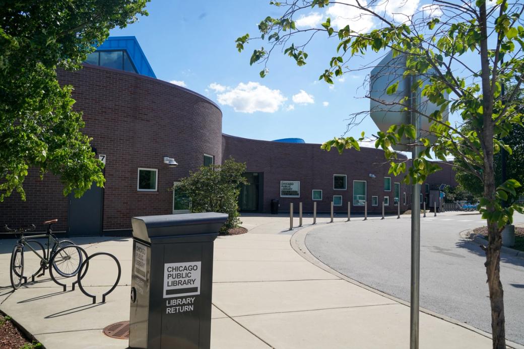 The exterior of the Altgeld Branch of the Chicago Public Library, a curved red brick building with blue skylight accents, on a sunny day. A book return drop box and bicycle rack are visible in the foreground.