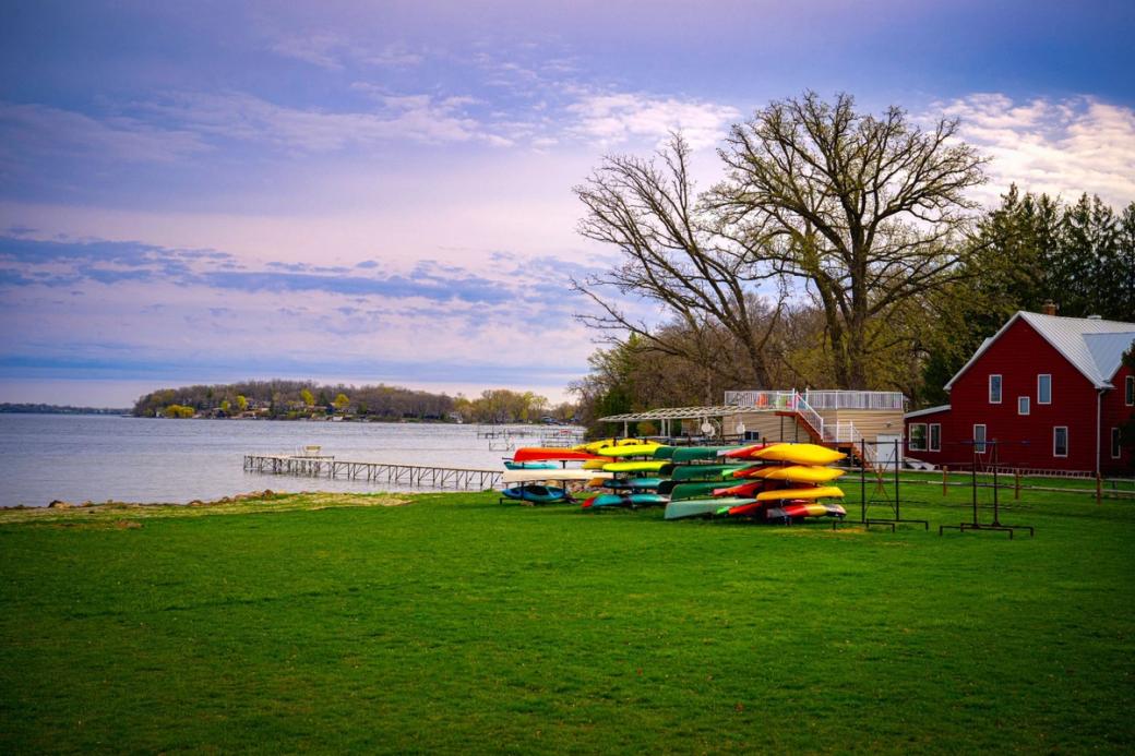 A red barn overlooks a lake, with yellow, green, and red surfboards lying on a large patch of green grass beside it.