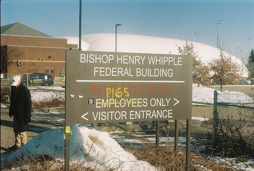 A photo of the sign in front of the Whipple Federal Building. Sign reads: "Bishop Henry Whipple Federal Building, Employees Only, Visitor Entrance." In yellow spray paint someone has written "PIGS"
