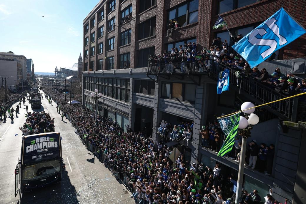 a photo of the Seattle Seahawks Championship Parade, with the float on the left carrying the team and fans and onlookers on the right. They are on the street and on balconies, one of which has a big number 12 flag)