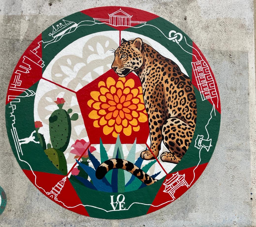A circular mural of a jaguar and a cactus, framed inside a red and green border. Along the border are Philly symbols including the Liberty Bell, Friendship Gate, and Rocky statue