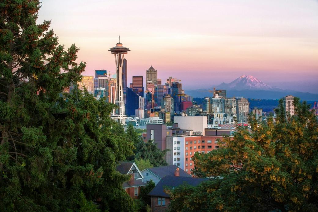 A sunset shot of Tahoma capped in snow with the Space Needle and the city skyline filled with apartment buildings on the left of the mountain.
