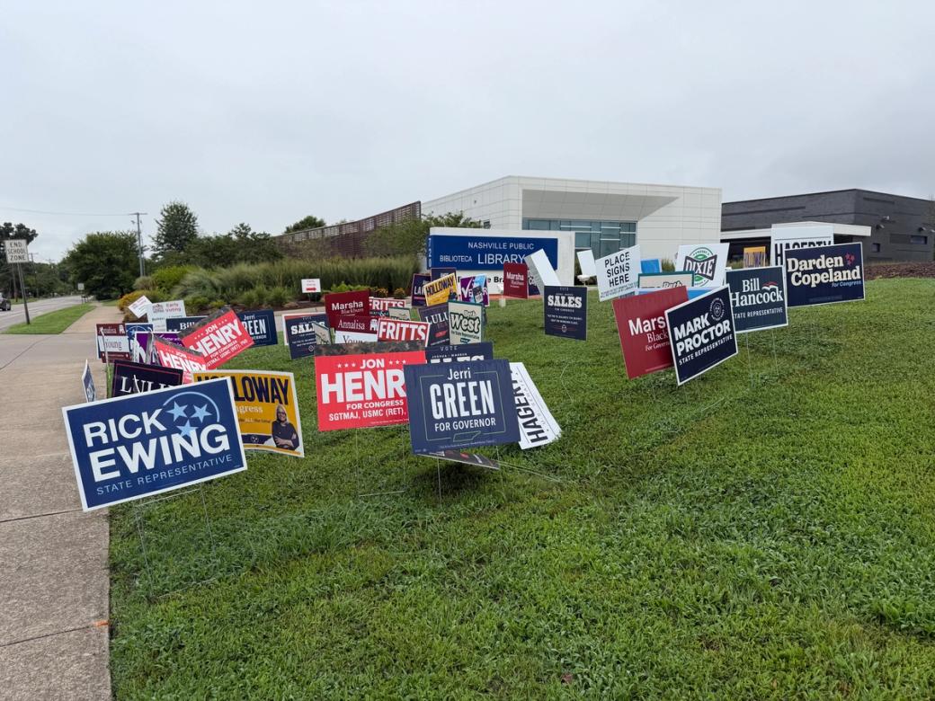 A collection of campaign sign on a grassy hill outside a white and gray building.