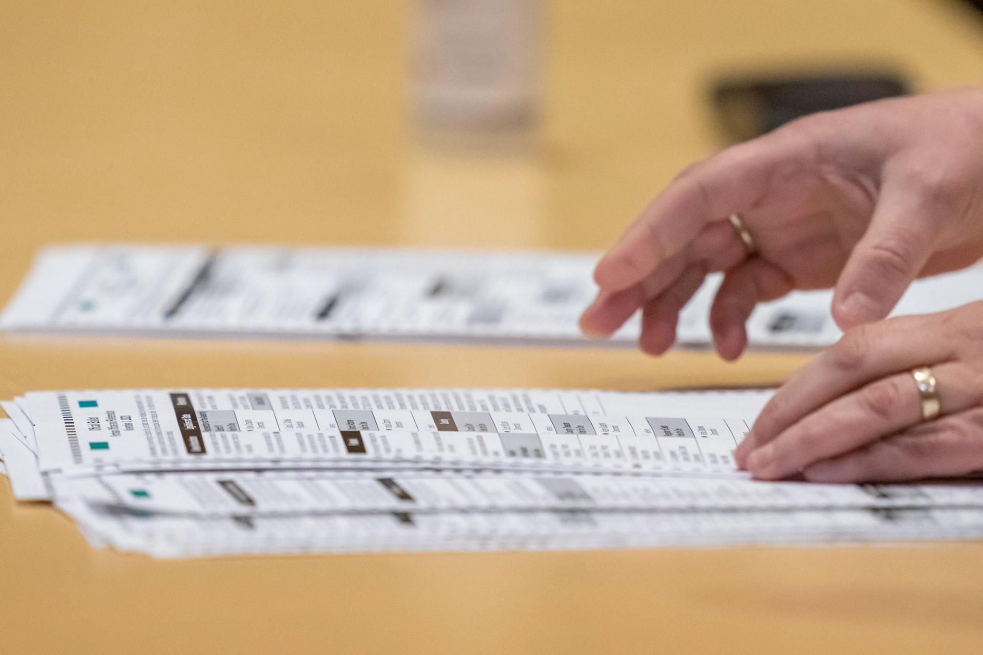 Two hands touch white paper ballots on a brown table.