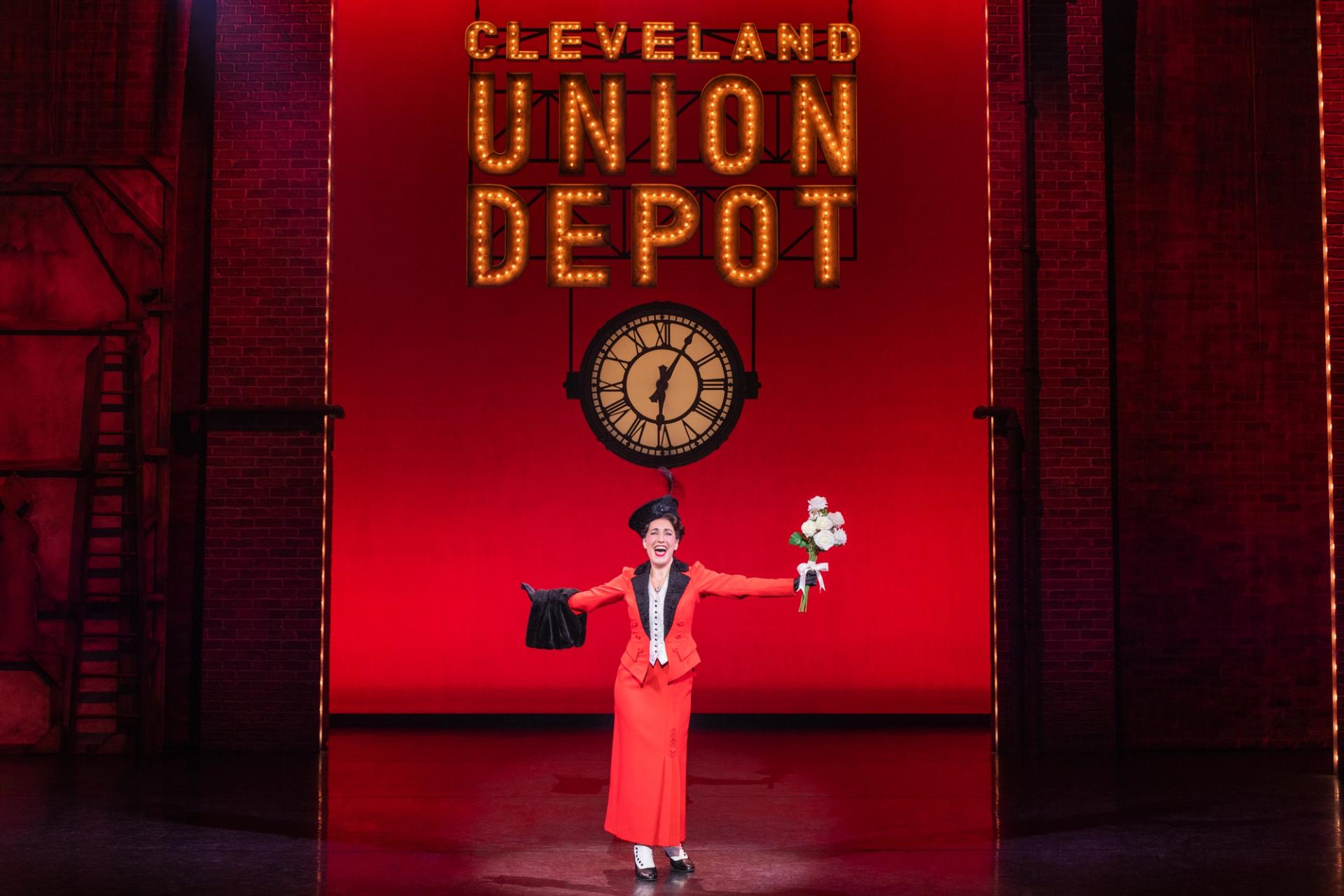 A woman in a red skirt and suit jacket sings on a stage.