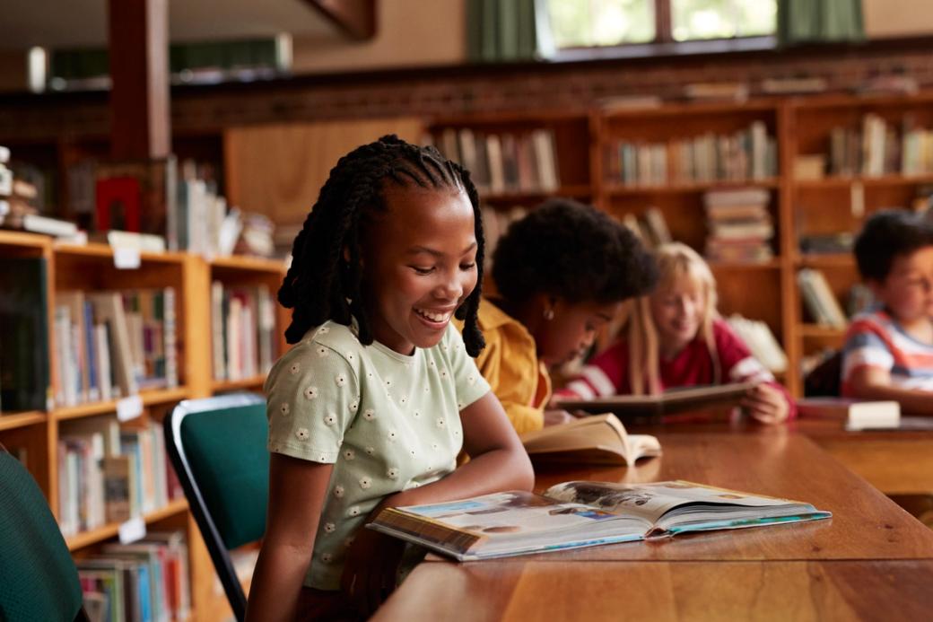 Four children sitting at a table reading, with bookshelves in the background.