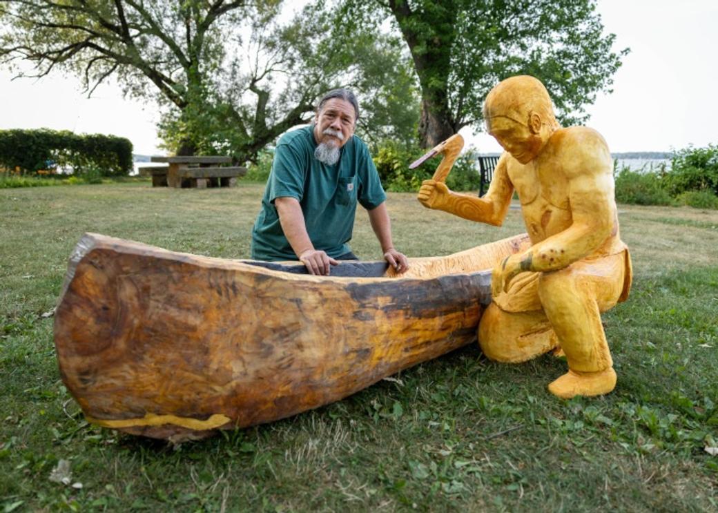 A man stands next to a piece of public art - a man in yellow shown carving a wooden canoe.