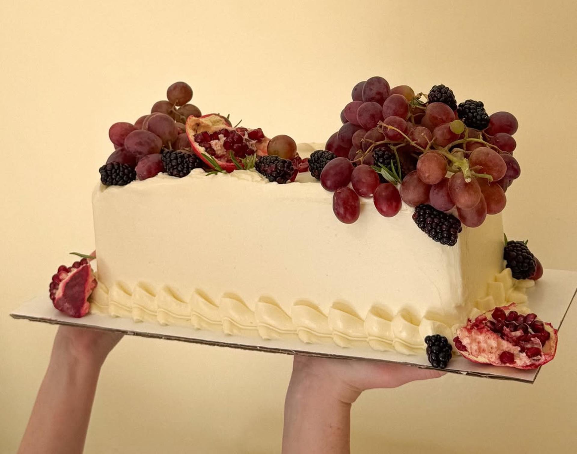 Person holding up rectangle cake decorated with grapes, pomegranate, and blackberries.