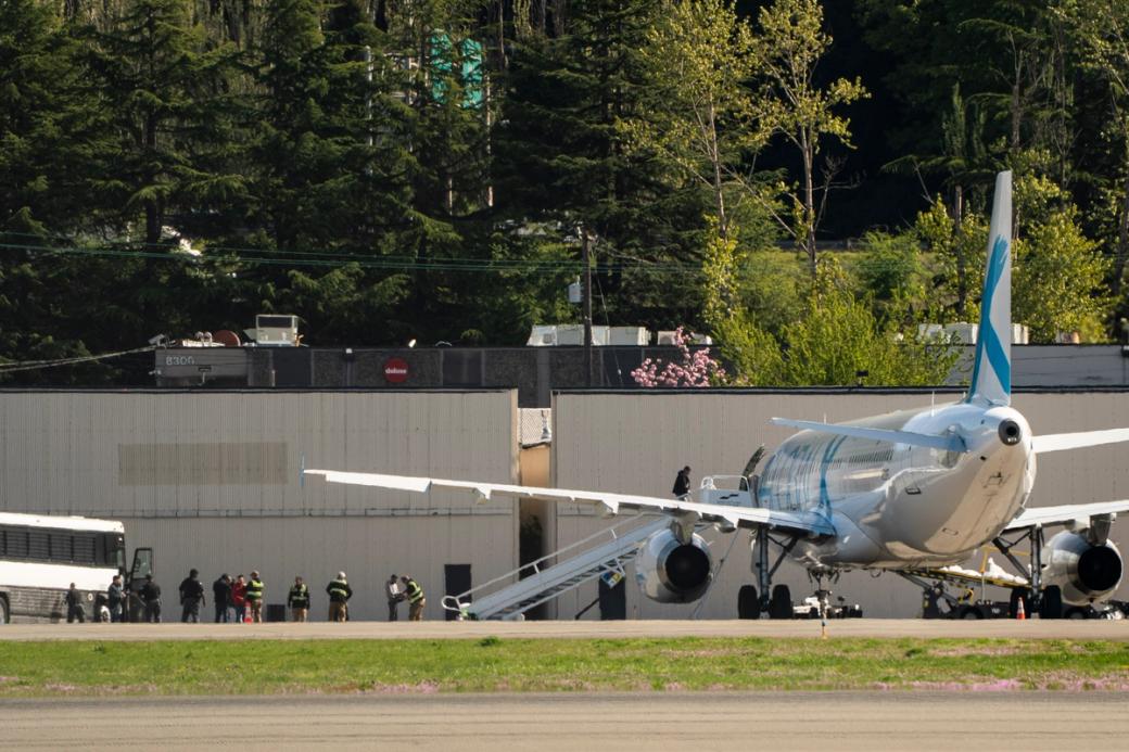 A plane sits on the tarmac at King County airport with a stairway down to the pavement. A line of people wait to board the plane to be transferred to detention facilities or deported out of the country