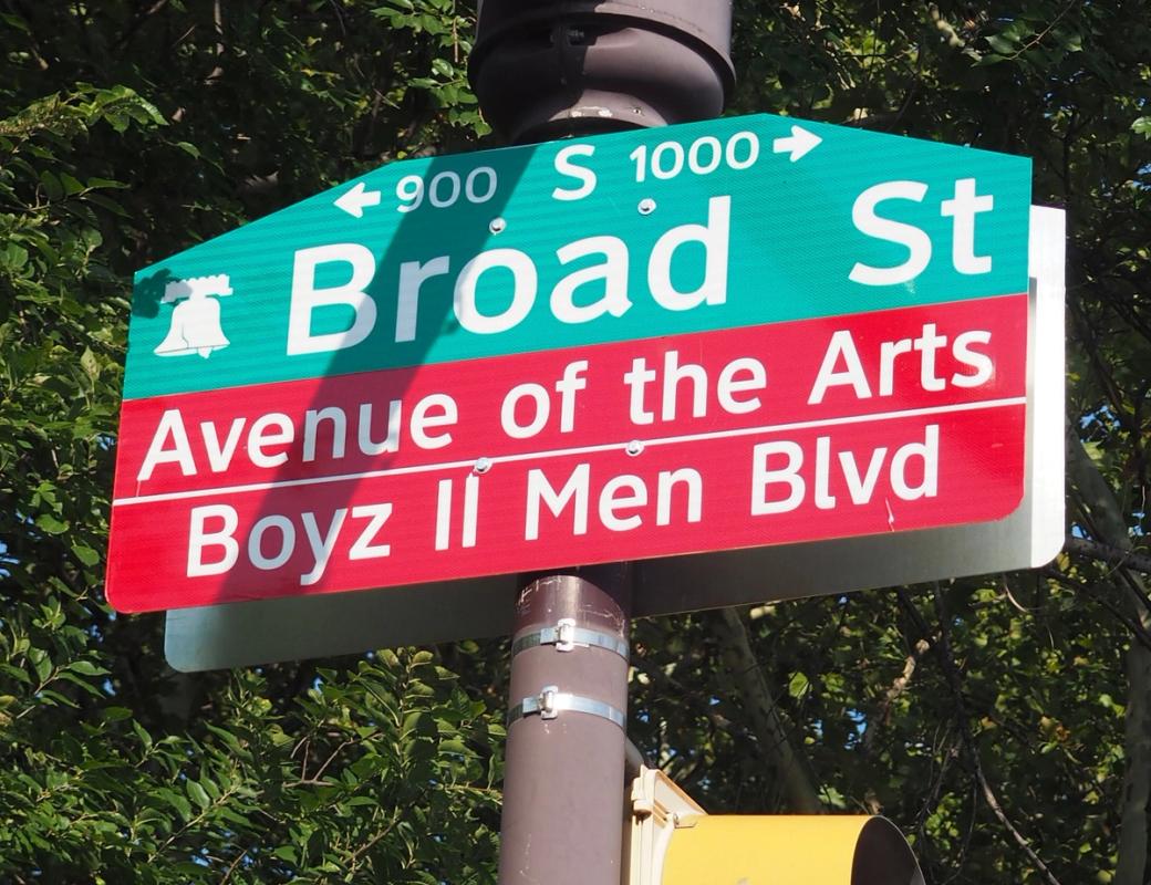 A green street sign pointing to the 900 and 1000 blocks of South Broad Street hangs on a pole. A red sign directly below the street name declares this section of the street is known as Avenue of the Arts and Boyz II Men Blvd. 