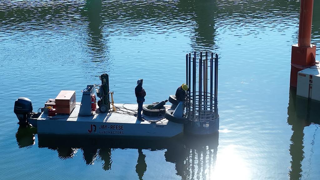 A person on a white, platform boat works on a bridge abutment.