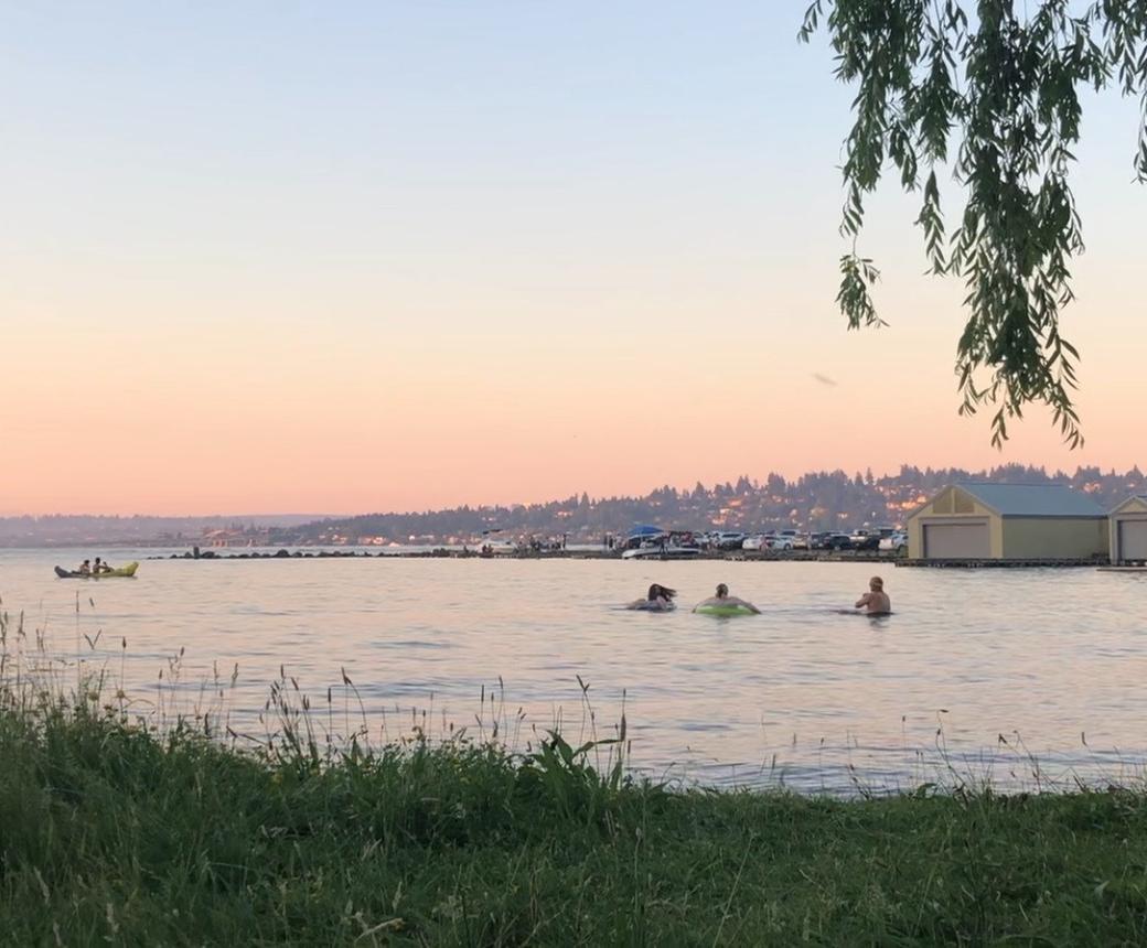 Three people float in Lake Washington along Seward Park, trying to beat the heat during the 2021 heatwave. The sky is a hazy pink and orange sunset in the background.