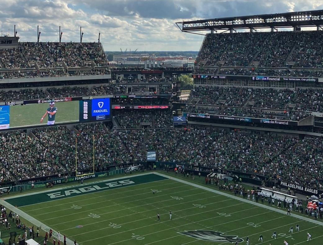 A view of a sold-out stadium at Lincoln Financial Field, with seats filled with fans and players on the field. The Eagles logo is visible at the center of the field, and the wordmark at an endzone. A player is projected on a screen inside the stadium.