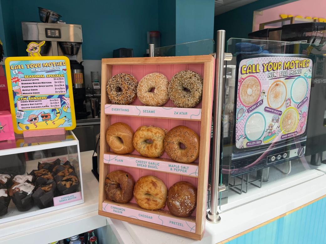 A bright pink, white, and blue display at Call Your Mother displays different bagels for sale, including everything, sesame, and za’atar flavors. A regular menu is on display, along with a fall menu. 