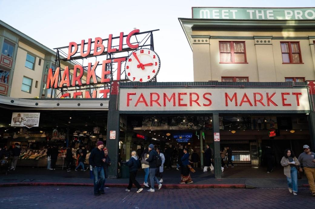 An image of Pike Place with shoppers and tourists checking out the stalls below the public market sign and farmers market