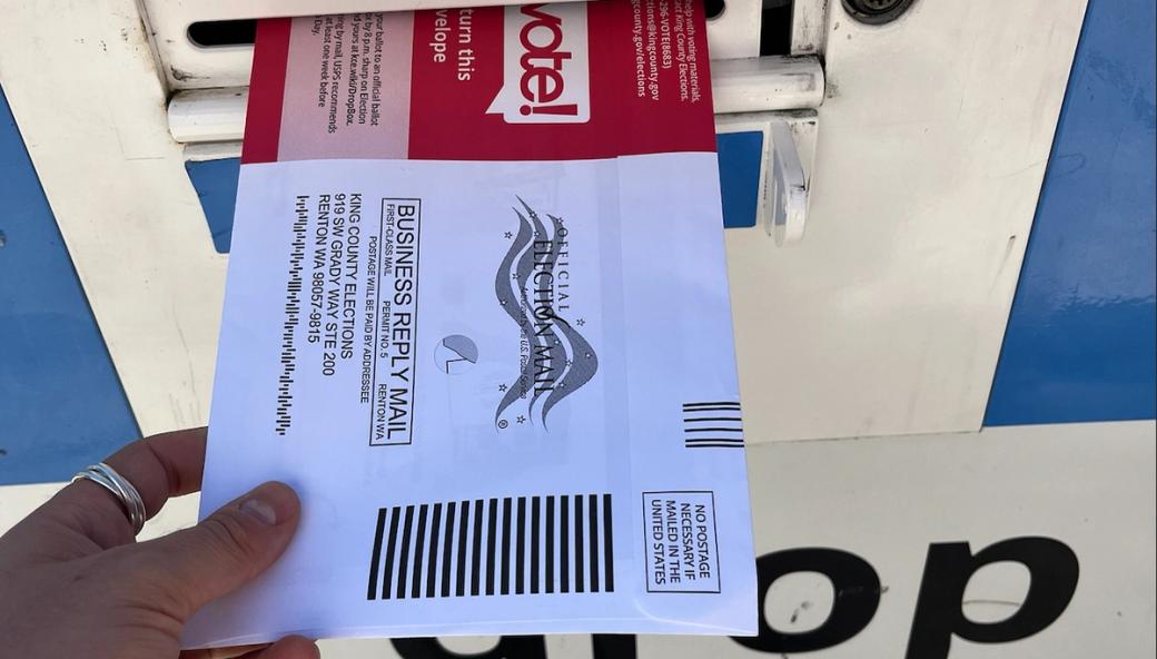 A hand pushes a King County Elections ballot envelope into a blue and white dropbox during the 2026 primary elections.