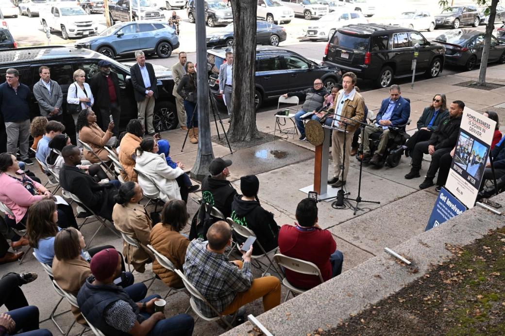 Mayor Mike Johnston at a podium in front of a small crowd on a sidewalk, with a sign next to him that reads "Homelessness is Down in Denver."