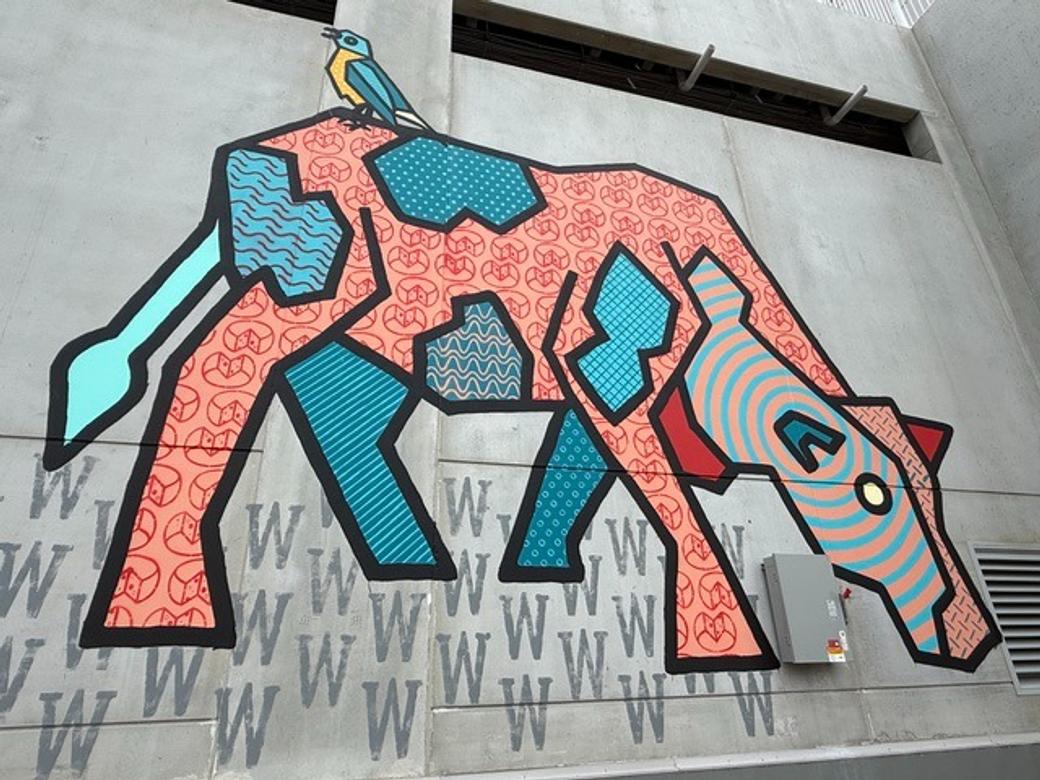 A mural on a building of a cow. The cow has patches of different shapes and colors - orange, turquoise.