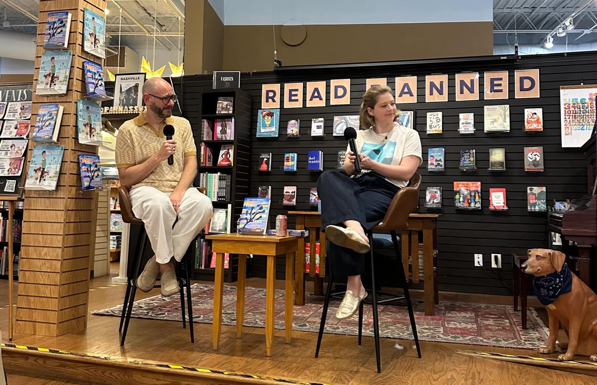 A man and woman sit on stools on the stage of a bookstore holding microphones. Behind them is a wall of books and a sign that says "Read Banned." A dog statue wearing a bandanna sits in the right hand corner of the photo.