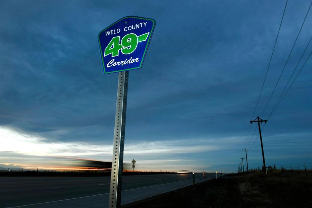 A blue-and-green traffic sign that says "Weld County 49 Corridor" is in the foreground, with a road, clouds, and power lines in the background.