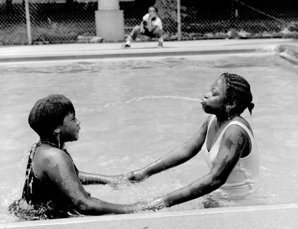 Two girls in a pool holding hands. There is a boy in the background crouching down. The girl on the right is spitting water at the other girl