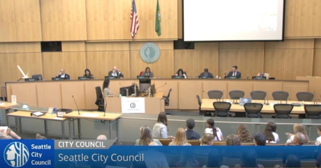 A screenshot of the Seattle Channel live stream of the August 11 city council meeting. Seven city council members sit behind their placards with the American and Washington flags behind them and the backs of many attendees in the audiences’ heads visible.