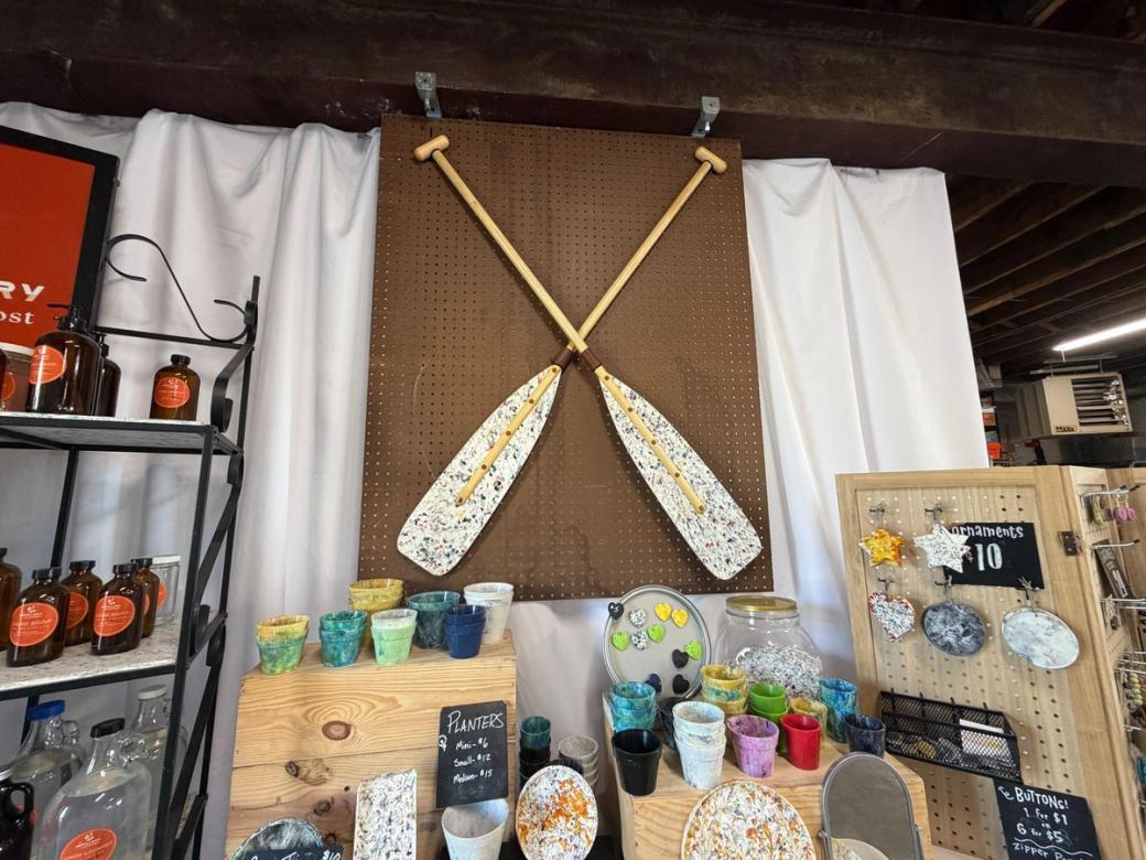 Two kayak paddles are mounted on the wall in an “X” formation above an assortment of flower pots and other wares for sale.
