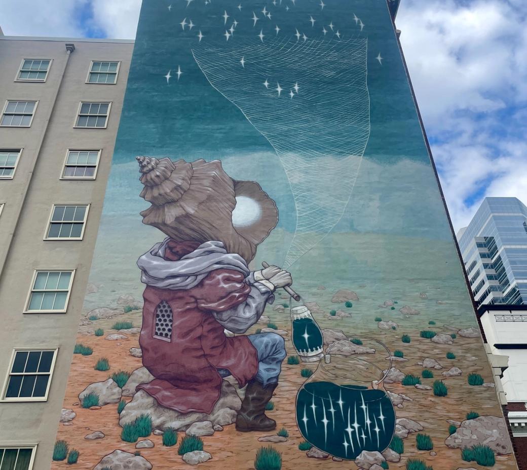 A mural covering the side of a tall building featuring a creature with a shell for a head sitting in the desert under a cloudy sky with stars.