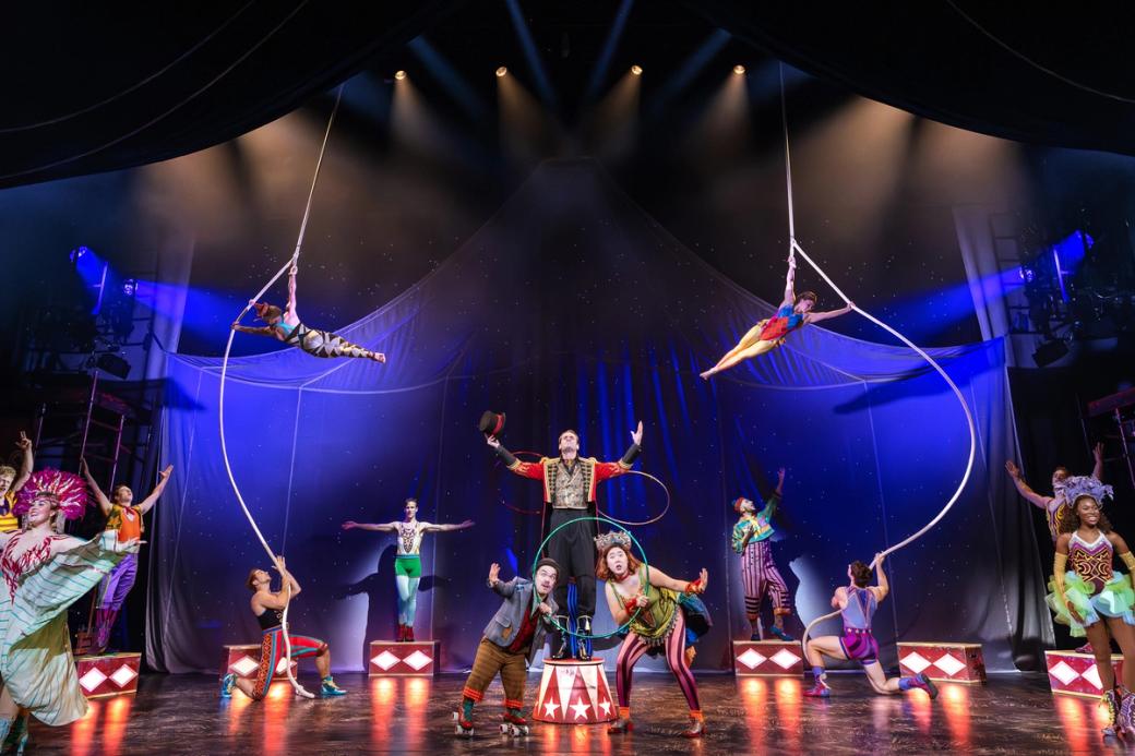 A vibrant circus stage with performers in colorful costumes. Aerialists swing gracefully above, while a ringmaster commands attention below, creating a lively scene.