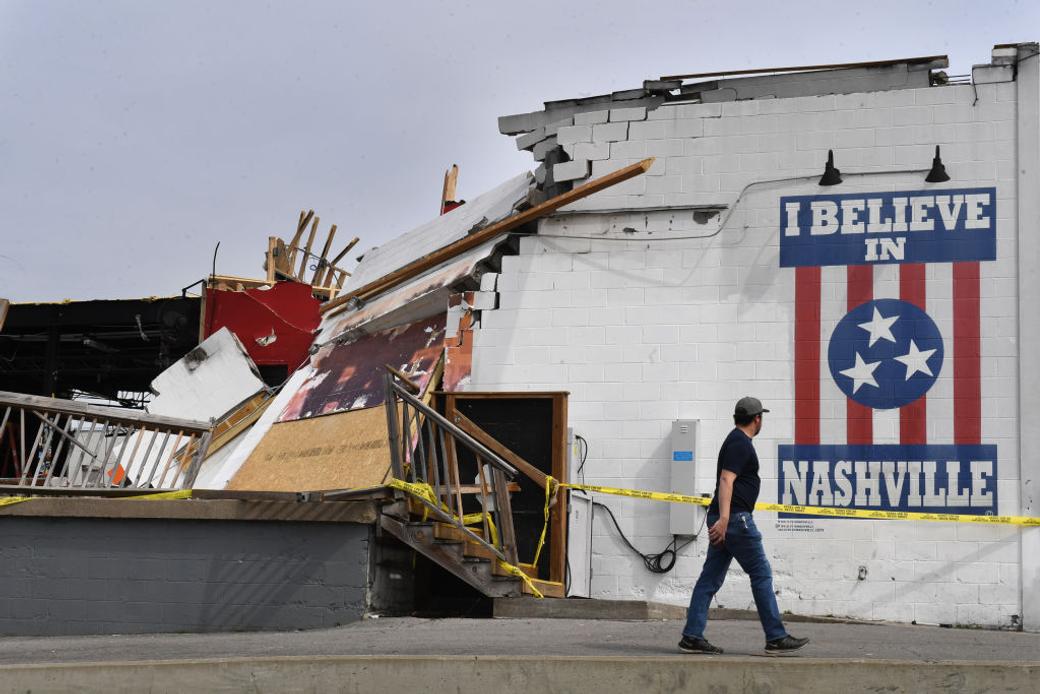 A white building with half of it caved in after a tornado. A mural with red and white stripes and a circle with Tennessee's three stars in the middle says I Believe in Nashville