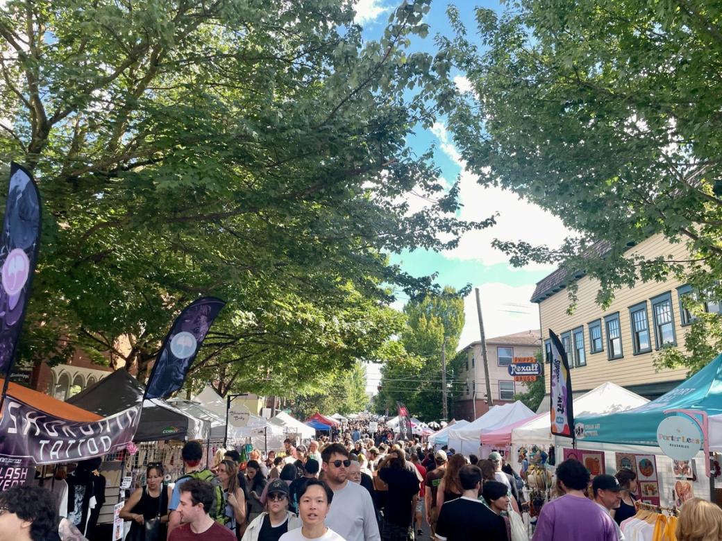 A large crowd of people fills a long street lined with trees and vendor booths on a sunny day.