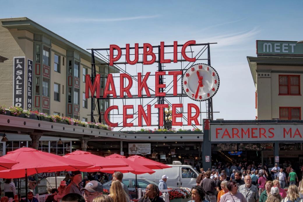 A photo of the red Public Market Center and clock at Pike Place market, with red umbrellas and pedestrians walking around the area