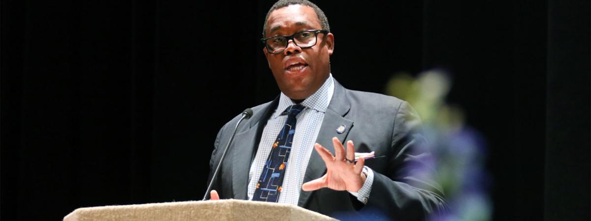 A dark-skinned man with black-rimmed glasses speaks at a pulpit or dais, one hand raised as if gesturing. He wears a gray suit and blue tie.