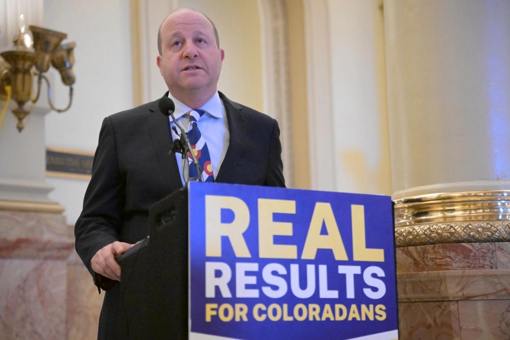 Gov. Jared Polis is standing behind a sign that reads "Real Results for Coloradans." He is in the Colorado State Capitol Building, next to a white and marble pillar.