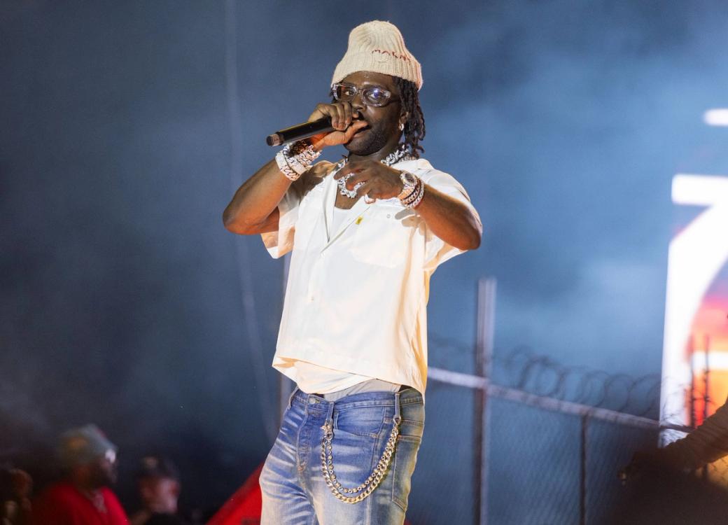 A Black man wearing jeans, a white shirt, beige knit hat, and jewelry holding a microphone. A fence and a few members of the audience can be seen in the background.