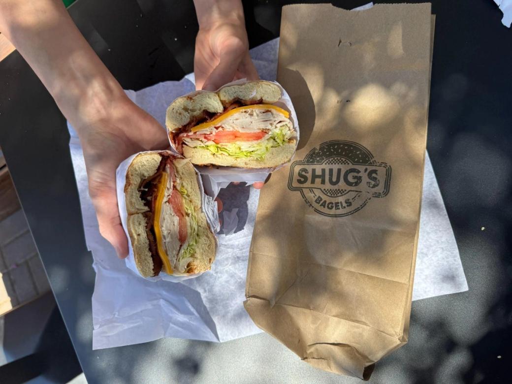 A person holding a bagel sandwich next to a brown paper bag that says "Shug's Bagels."