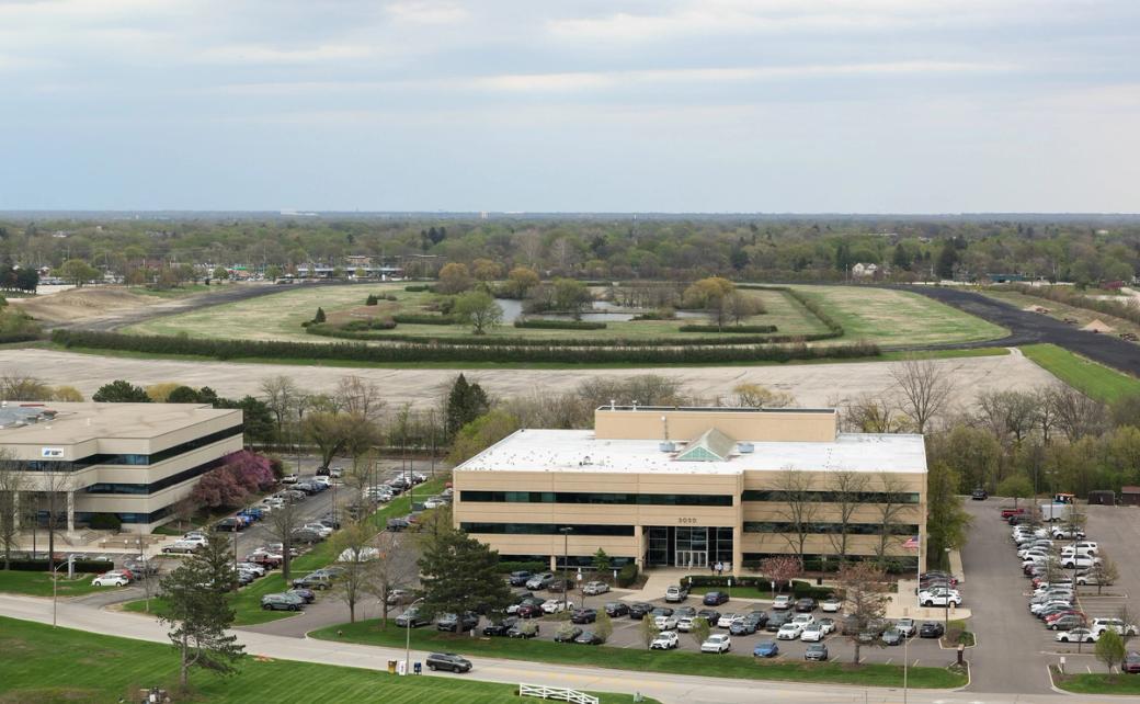 An aerial shot of an office building with a parking lot containing cars. Behind the building there is a large green park with trees and a lake in the middle